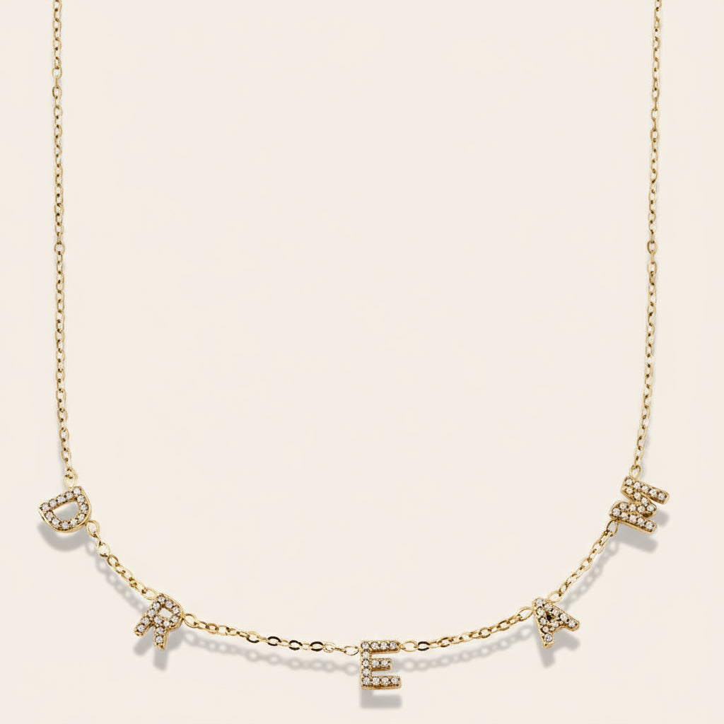 A delicate gold necklace with the word "DREAM" spelled out in individual letters, each letter adorned with a small sparkling crystal.